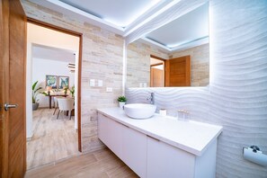 A modern bathroom with a large mirror, a white sink, and a wooden door. - Cana Brava Residences Rental Apartment  (Punta Cana)