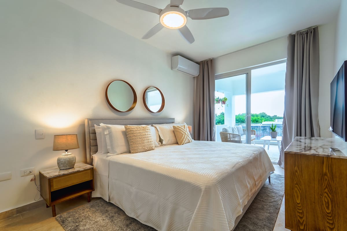 A bedroom with a bed, nightstand, lamp, ceiling fan, and a view of the outdoors.