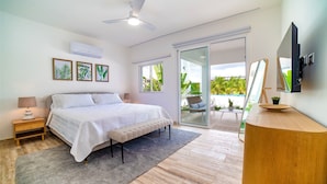 A bedroom with a bed, bedside tables, a ceiling fan, an air conditioner, a sliding glass door to a balcony, and a TV mounted on the wall. - Cana Brava Residences Rental Apartment  (Punta Cana)