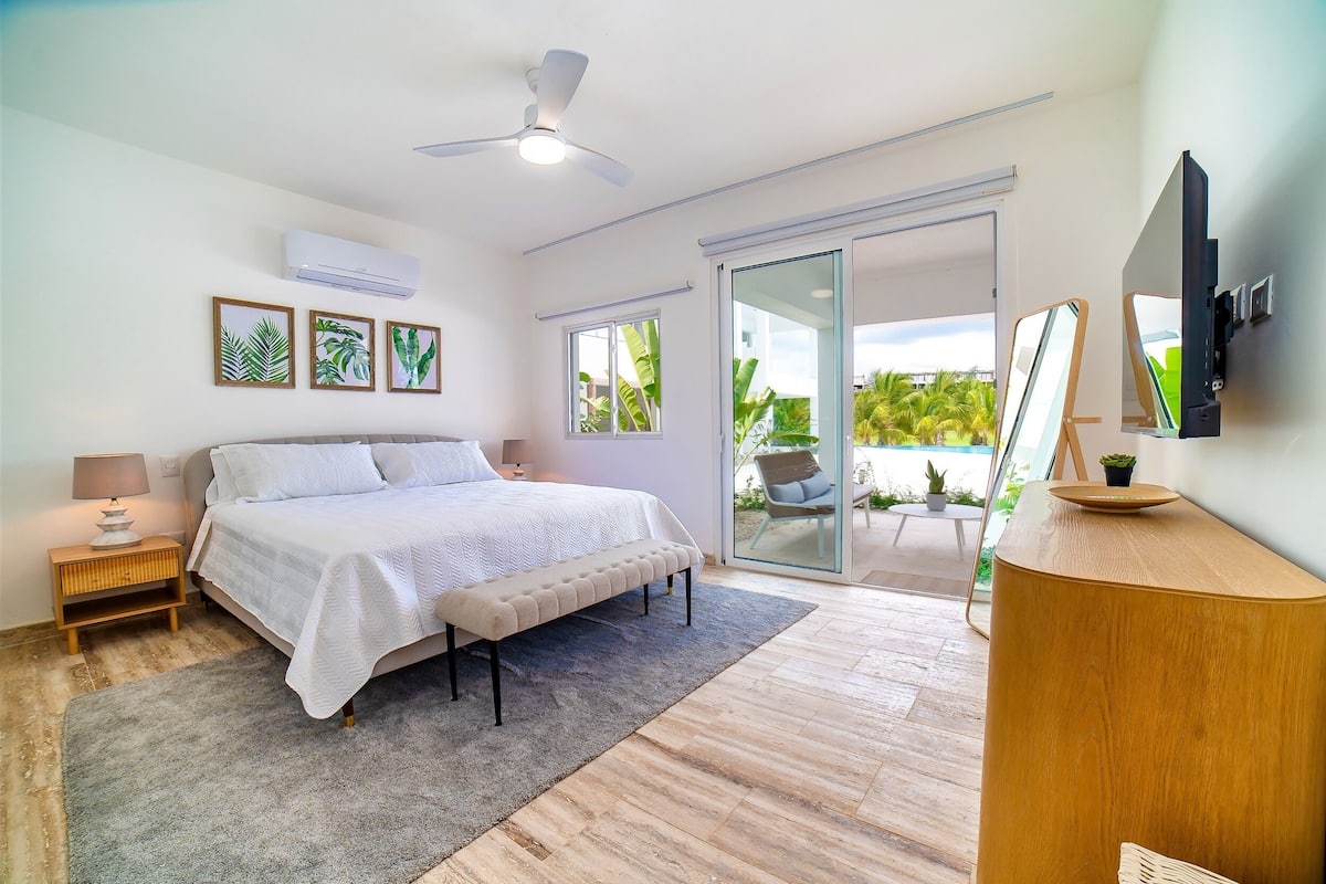 A bedroom with a bed, bedside tables, a ceiling fan, an air conditioner, a sliding glass door to a balcony, and a TV mounted on the wall.