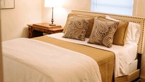 A bedroom with a bed, a nightstand, a lamp, a ceiling fan, and a window with blinds. - Boutique Beeville Cottage (Beeville)