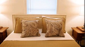 A bedroom with a headboard, two patterned pillows, a bedspread, and a window with blinds. - Boutique Beeville Cottage (Beeville)