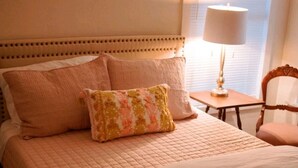 A neatly made bed with a quilted cover and two pillows, a bedside lamp, and a chair in a room with a window and blinds. - Boutique Beeville Cottage (Beeville)