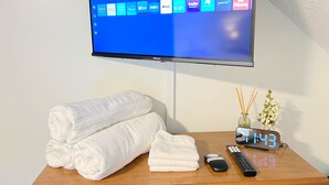 A flat-screen TV mounted on a wall displaying various streaming apps, a wooden side table with folded towels, a remote control, and a digital clock showing 1:43.