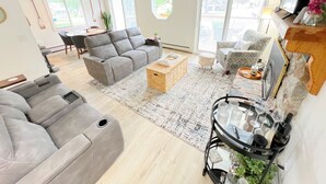 A spacious living area with a grey sofa, a patterned armchair, and a coffee table. There is a dining area in the background with a table and chairs.