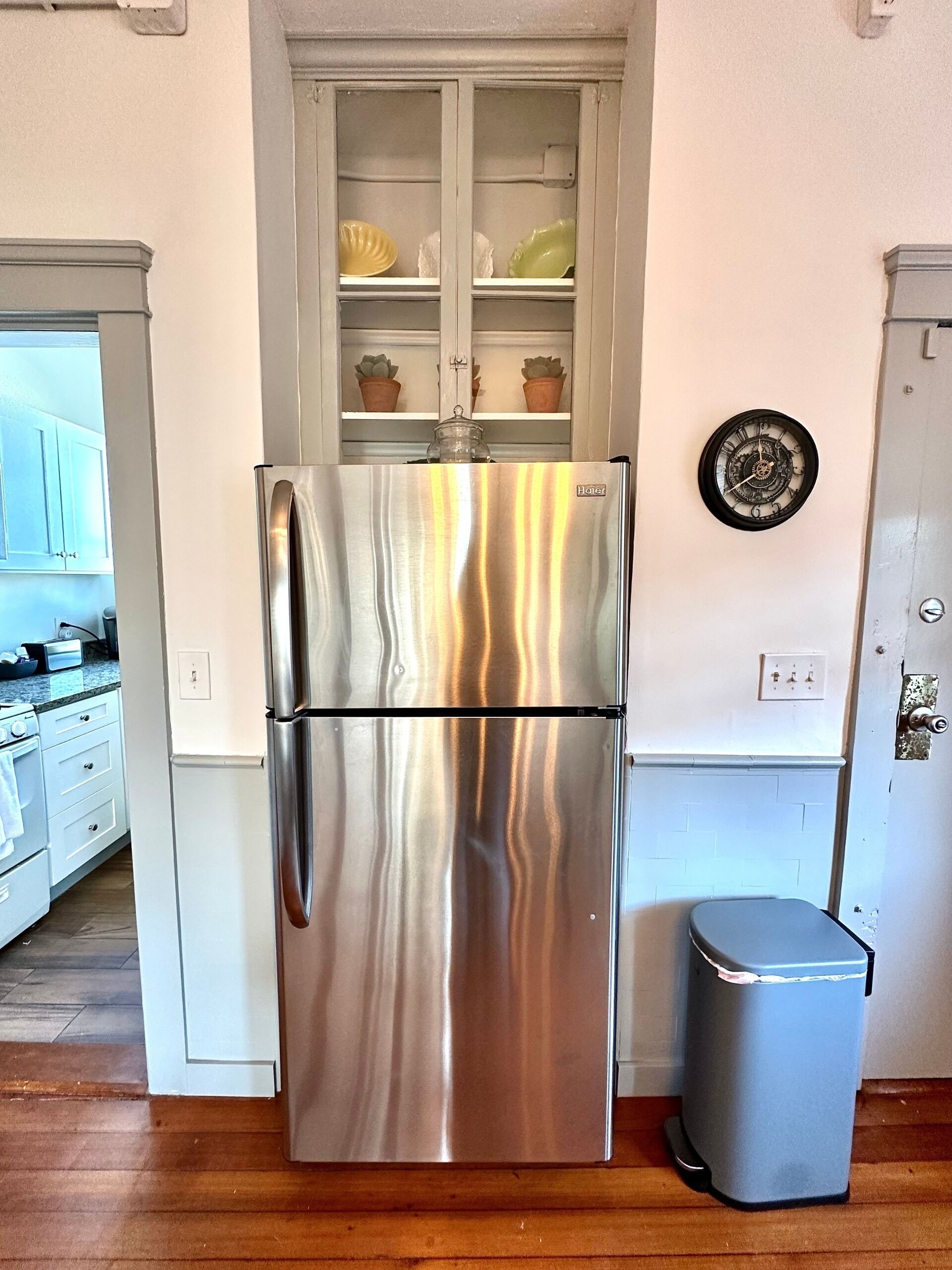 A stainless steel refrigerator in a kitchen with a cabinet above it containing decorative items.