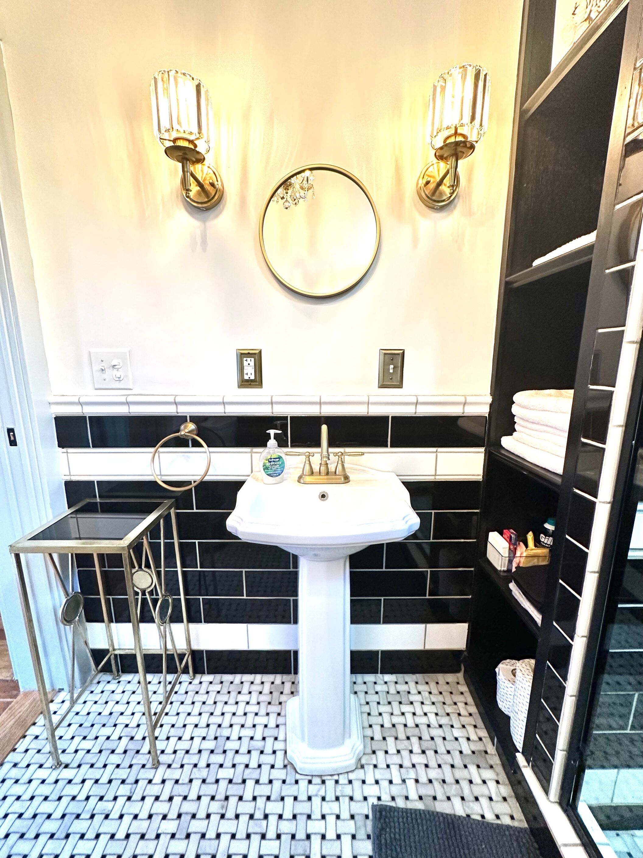 A bathroom with a white pedestal sink, black and white tiled backsplash, two wall-mounted lights, a round mirror, and a black shelving unit with towels.