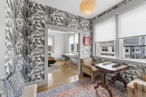 A room with a chessboard, a striped armchair, a patterned rug, and a view of buildings through large windows. - 3BR Luxe Penthouse Federal Hill/Downtown
Perfect homebase for FIFA World Cup2026 (Providence)