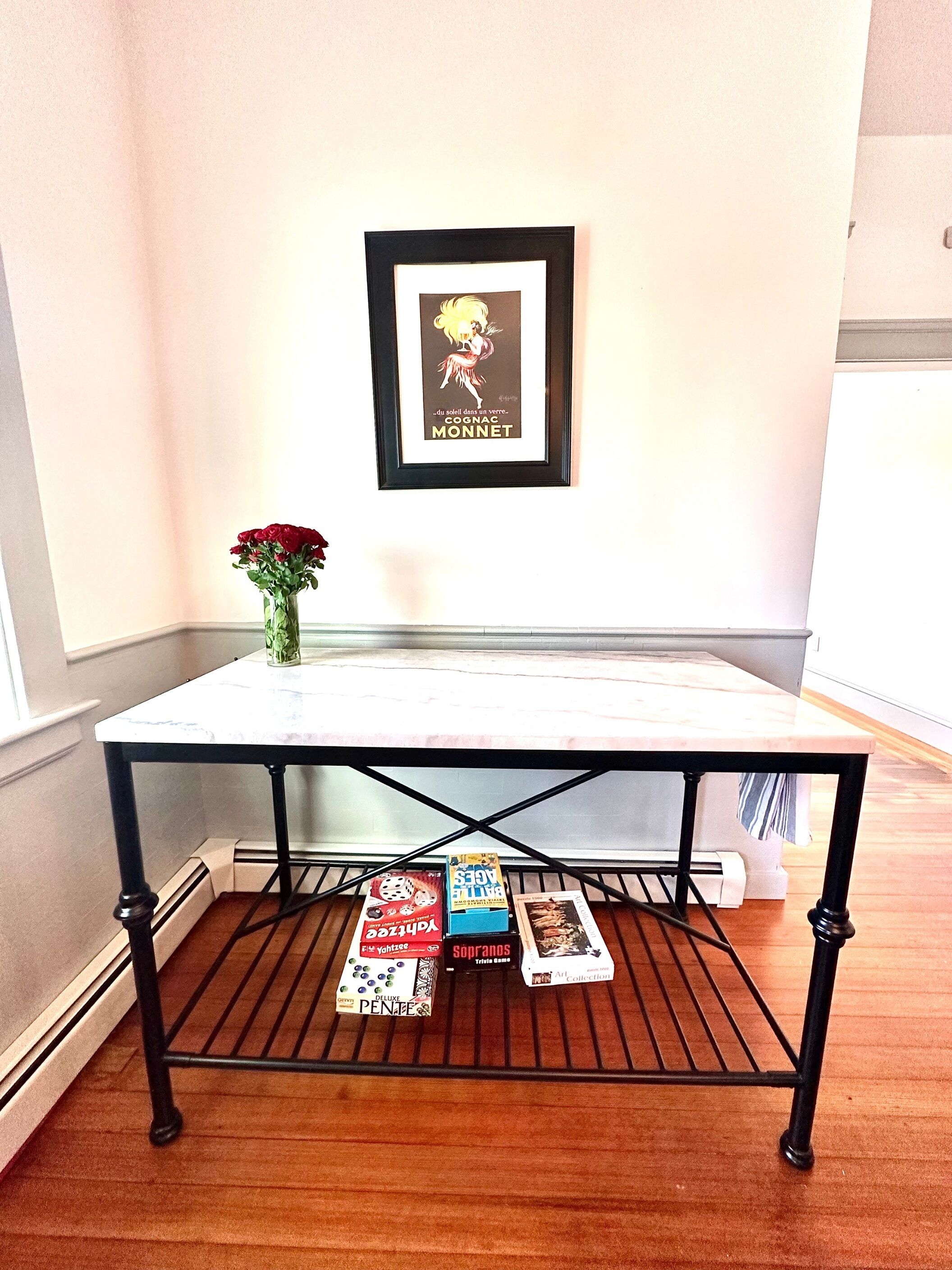 Uma pequena mesa com tampo branco e estrutura de metal preto, colocada em um canto de um quarto com parede branca e piso de madeira.