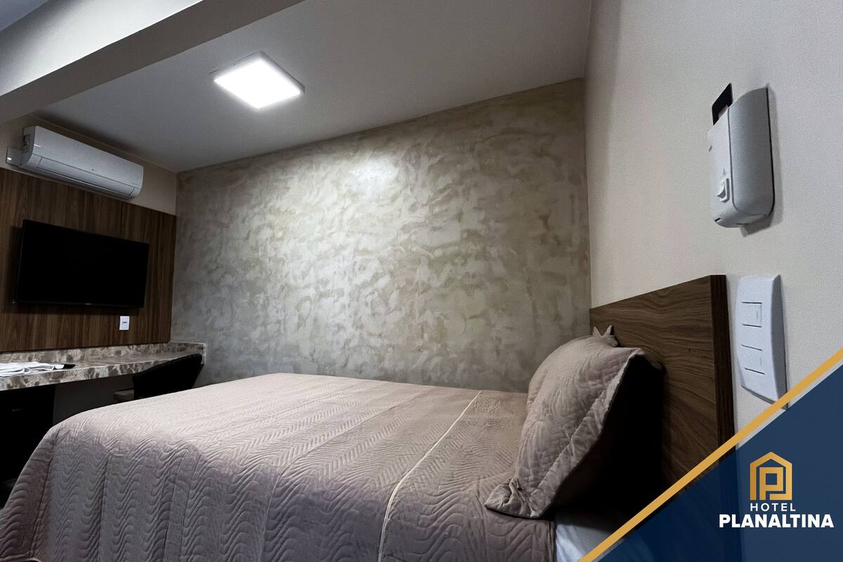 Habitación de hotel con cama, televisor montado, tocador con espejo y aire acondicionado de pared.