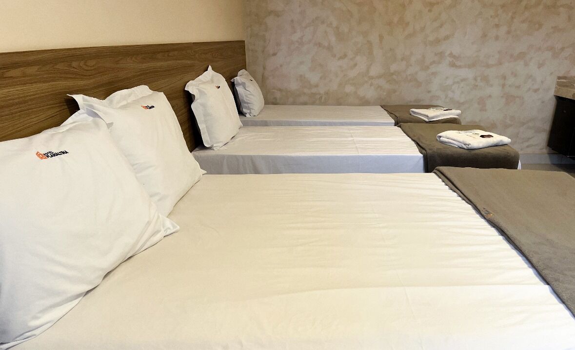A hotel room with three beds, each with white linens and pillows. There is a small table with a towel and a remote control.