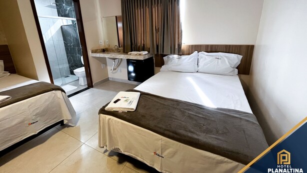 A hotel room with a bed, a bedside table, a mirror, a window with curtains, and an air conditioning unit. - Planaltina (Planaltina)