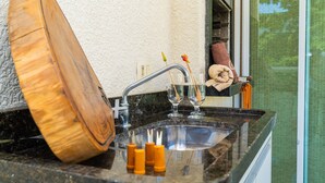 A kitchen with a granite countertop, a wooden cutting board, a glass of water, and a towel hanging on the wall. - Elegante Apartamento em Enseada no Guarujá (Guarujá)