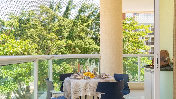 A balcony with a table set for dining, chairs, and a view of trees and buildings. - Elegante Apartamento em Enseada no Guarujá (Guarujá)