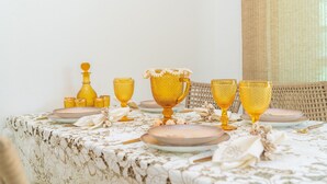 A dining table set with golden glassware and plates, a wicker chair, and a patterned tablecloth. - Elegante Apartamento em Enseada no Guarujá (Guarujá)