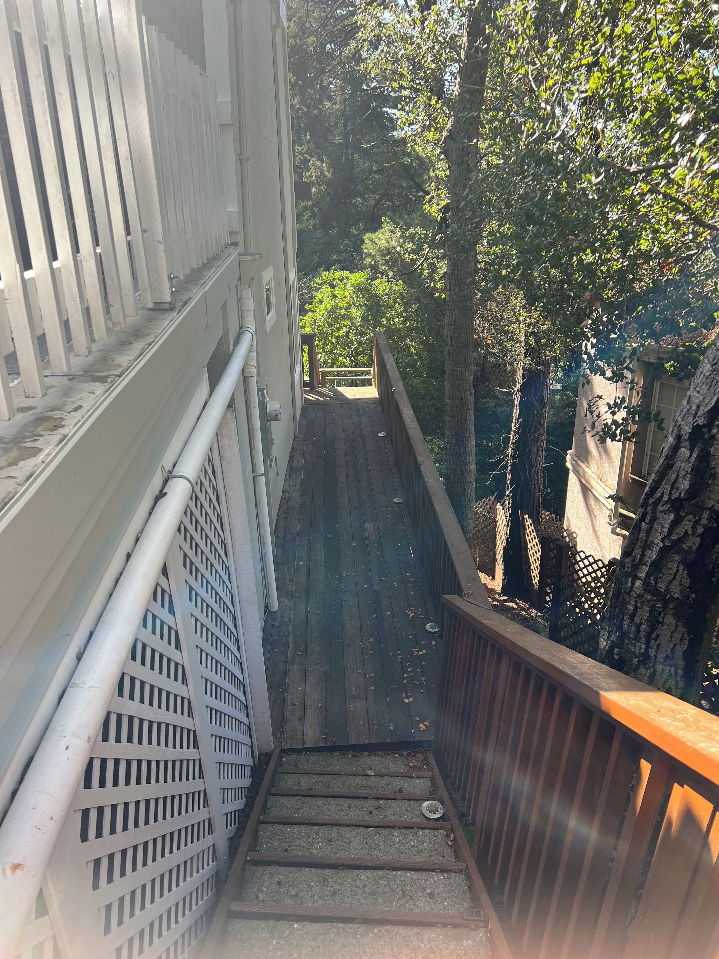 A wooden staircase leading to a balcony with a metal railing, surrounded by trees and a building facade.