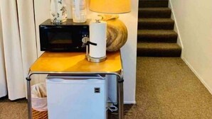Ein kleines Hotelzimmer mit Schreibtisch, Lampe, Kaffeemaschine, Minikühlschrank und Ablagefläche mit Tassen und Wasserkocher.