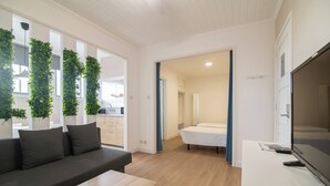 A modern living room with a sofa, coffee table, and a TV on a stand. There is a kitchen area visible through an open door. - Livramento Residence (Ponta Delgada)