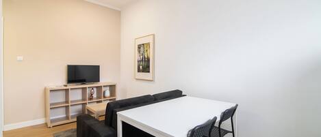 Een moderne woonkamer met een witte tafel, zwarte stoelen, een zwarte bank, een televisie op een houten dressoir en een ingelijst schilderij aan de muur.