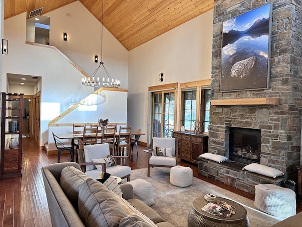 A spacious living room with a stone fireplace, a high ceiling, and a dining area with a staircase.