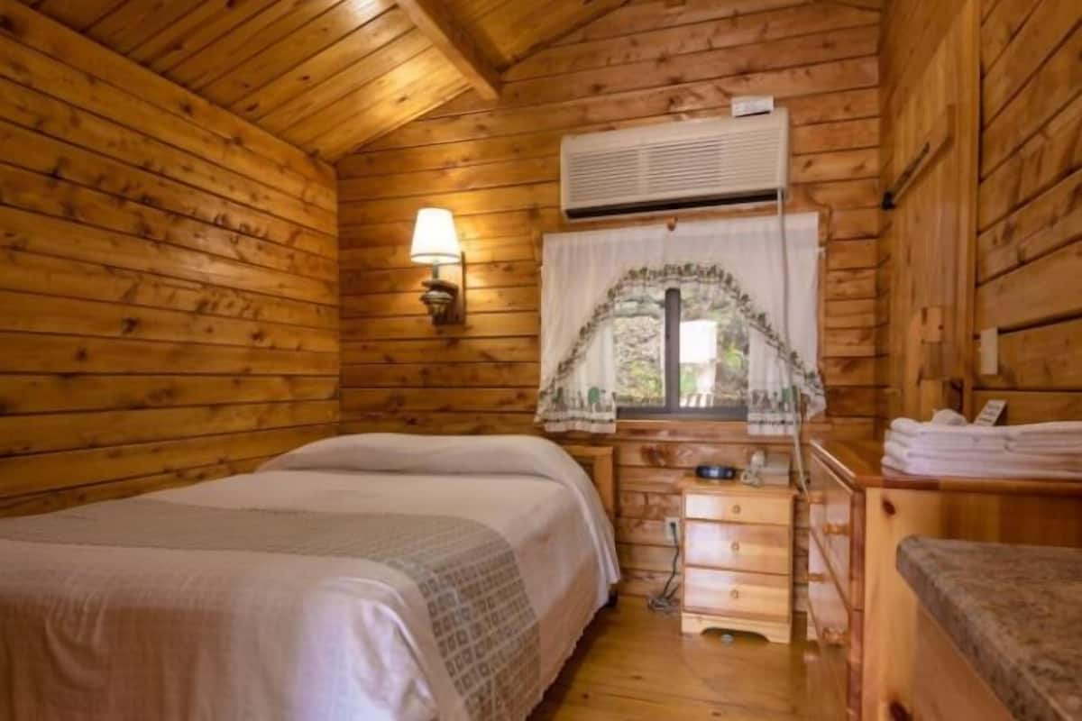 A wooden cabin room with two beds, a nightstand, a lamp, and a window with curtains.