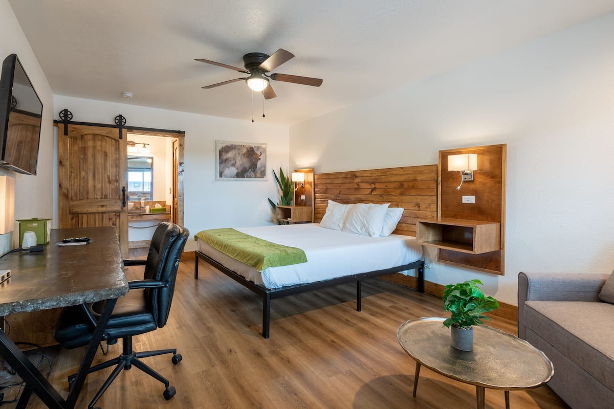 A compact room with a bed, a desk, a chair, a ceiling fan, a small table with a plant, and a wooden cabinet.