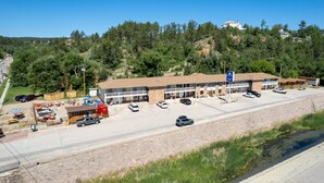 A motel with multiple parking spaces, a small outdoor seating area, and a view of a forested hillside. - Springs Lodge & Cafe (Hot Springs)