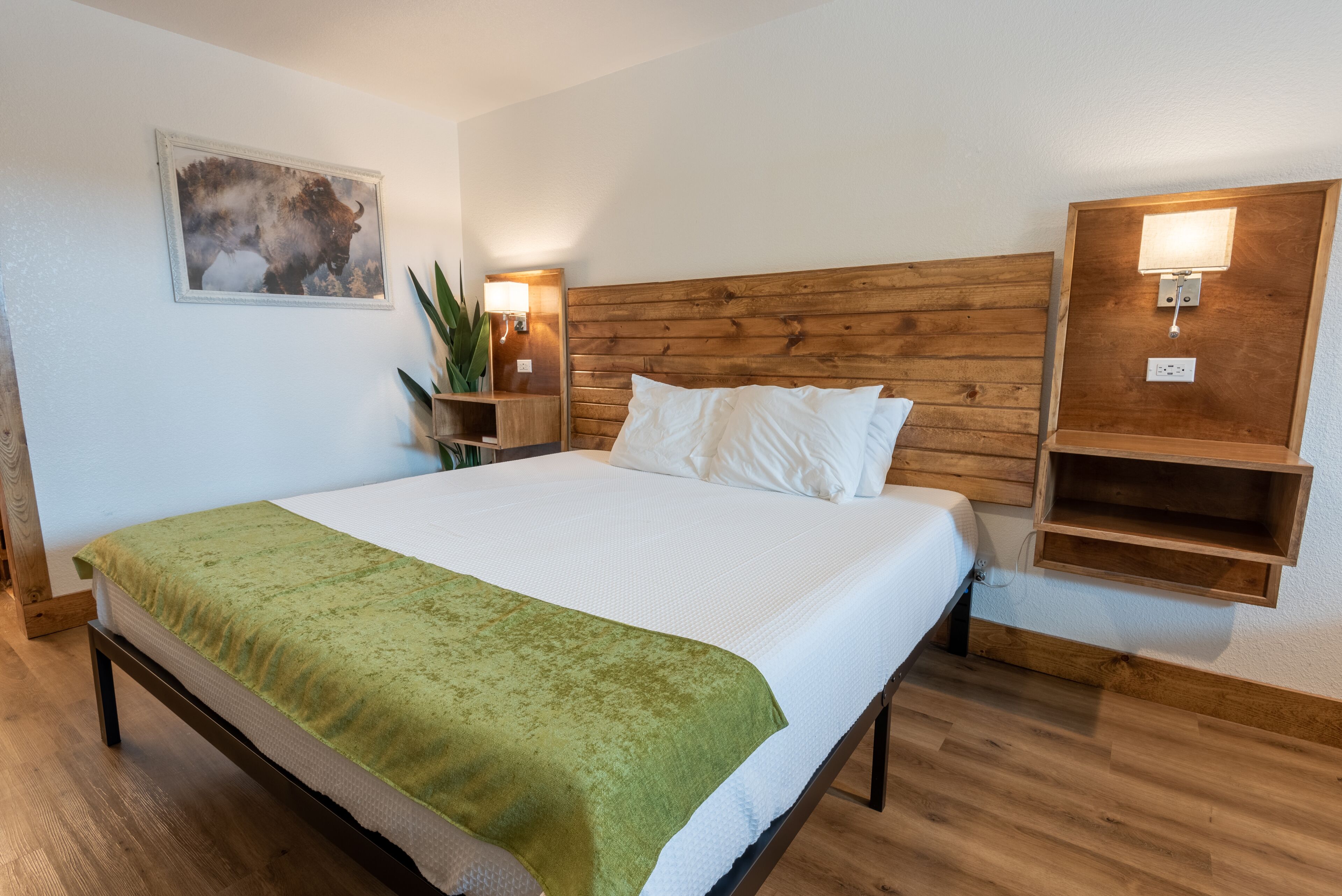 A neatly arranged hotel room with a large bed, wooden headboard, bedside lamps, a wall-mounted shelf, and a framed picture.