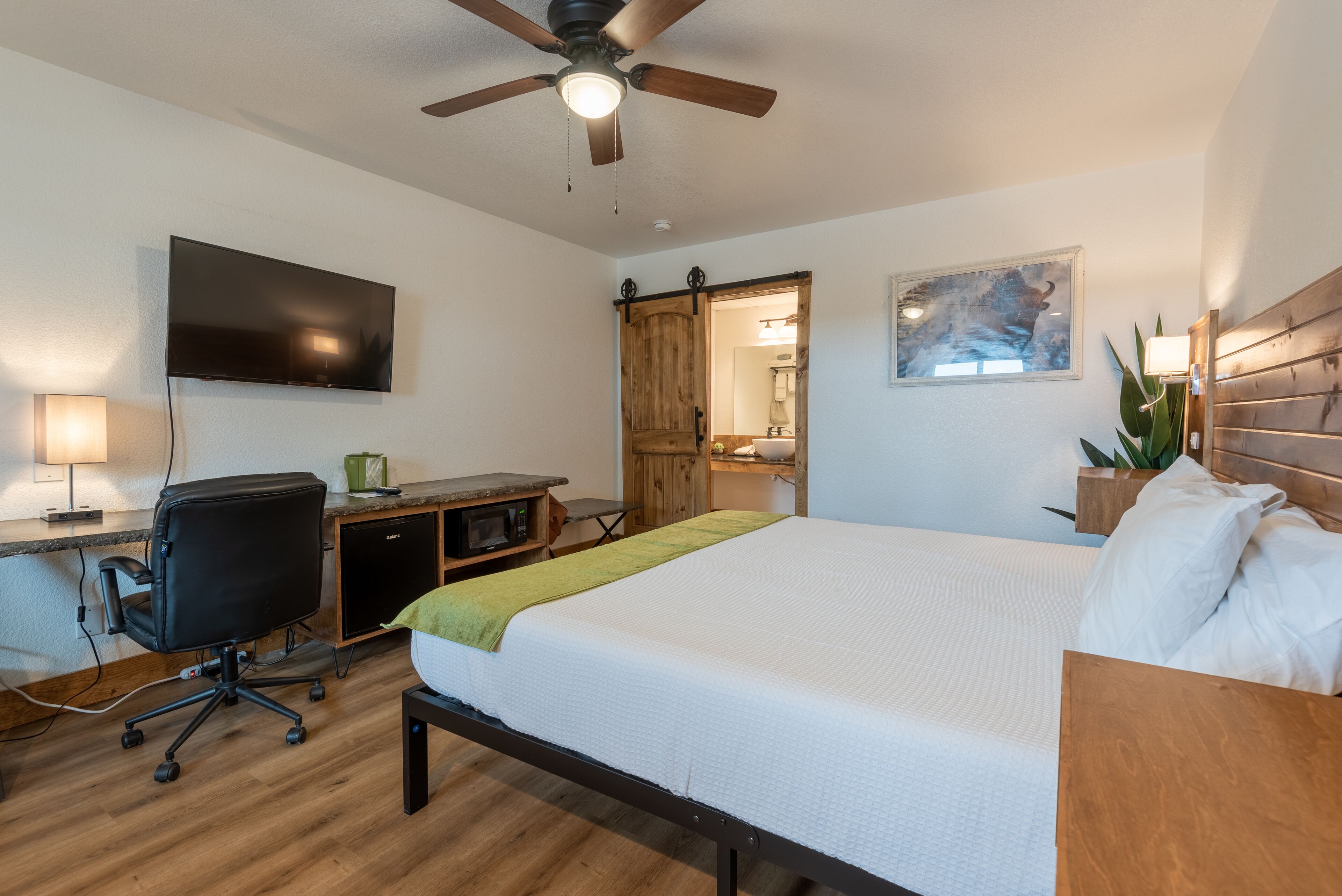 A hotel room with a bed, desk, chair, television, ceiling fan, and a wooden wardrobe.