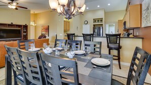 A dining area with a large table set for six, surrounded by wooden chairs. A chandelier hangs above the table, and a television is mounted on the wall to the left. - Resort Perks, Patio & Gas Grill: Indio Retreat! (Indio)