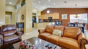 A living room with a brown leather sofa, a coffee table with remote controls, and a dining area with a chandelier. - Resort Perks, Patio & Gas Grill: Indio Retreat! (Indio)