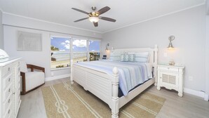Une chambre avec une grande fenêtre, un lit avec une literie à rayures, un ventilateur de plafond et une table de chevet avec une lampe.