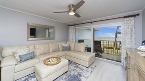 Un salon avec un canapé d’angle beige, un ventilateur de plafond, un grand miroir et une vue sur la plage, visible à travers une porte-fenêtre coulissante.