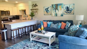 A modern living room with a blue sofa, colorful cushions, a coffee table, and a kitchen area with white cabinets and a microwave.
