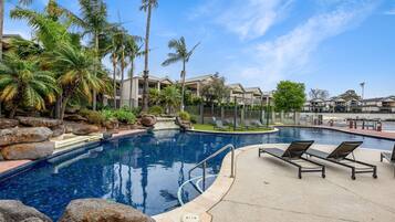 A swimming pool with lounge chairs, surrounded by palm trees and a building complex.