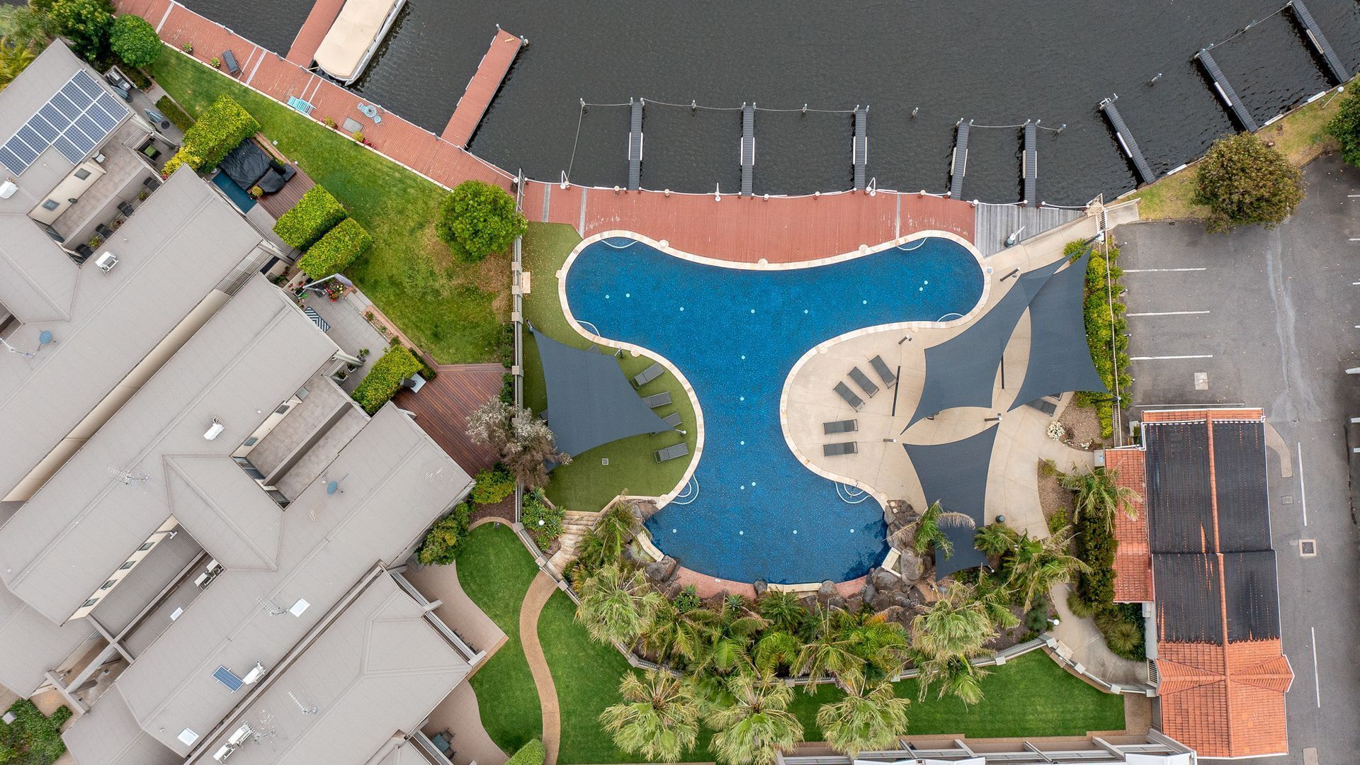Vue aérienne d’un quartier résidentiel avec une piscine en forme de guitare, entourée de verdure et jouxtant un parking.