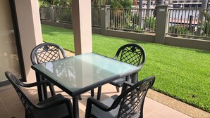 Une table à plateau en verre, entourée de quatre chaises noires, se trouve dans un espace patio extérieur offrant une vue sur une pelouse bien entretenue et une clôture en métal.