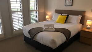 Un lit bien fait, avec du linge de lit blanc, un plaid marron et un coussin jaune. Deux lampes de chevet sont posées sur des tables de chevet en bois.