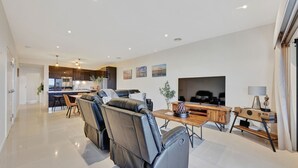 Een moderne woonkamer met een bank, salontafel en televisie. Achtergrond toont een keuken met een eethoek.