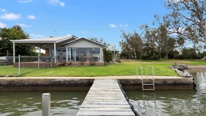 Une maison au bord du lac, avec un quai donnant sur l’eau, une terrasse couverte et une pelouse verdoyante.