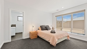 Een slaapkamer met een groot raam, een bed met een beige deken, een nachtkastje met een lamp en een houten dressoir.