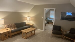Un espacio habitable estilo loft acogedor, con techo alto, una mesa de centro de madera, un sofá y una silla. Se ve una pequeña mesa con una lámpara, y una cama con ventana al fondo.