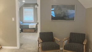 Una habitación con piso alfombrado, dos sillones, un cuadro en la pared y un acceso a un dormitorio con cama y ventana.