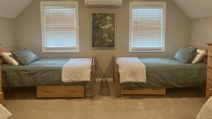 Un dormitorio con dos camas individuales, cada una con estructura de madera y colcha verde. Hay dos ventanas con persianas, un equipo de aire acondicionado sobre la ventana izquierda y un cuadro en la pared.