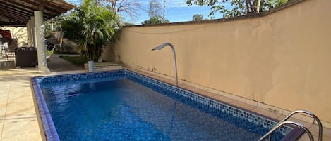 A rectangular swimming pool with a tiled edge, surrounded by a beige wall and a tiled patio area.