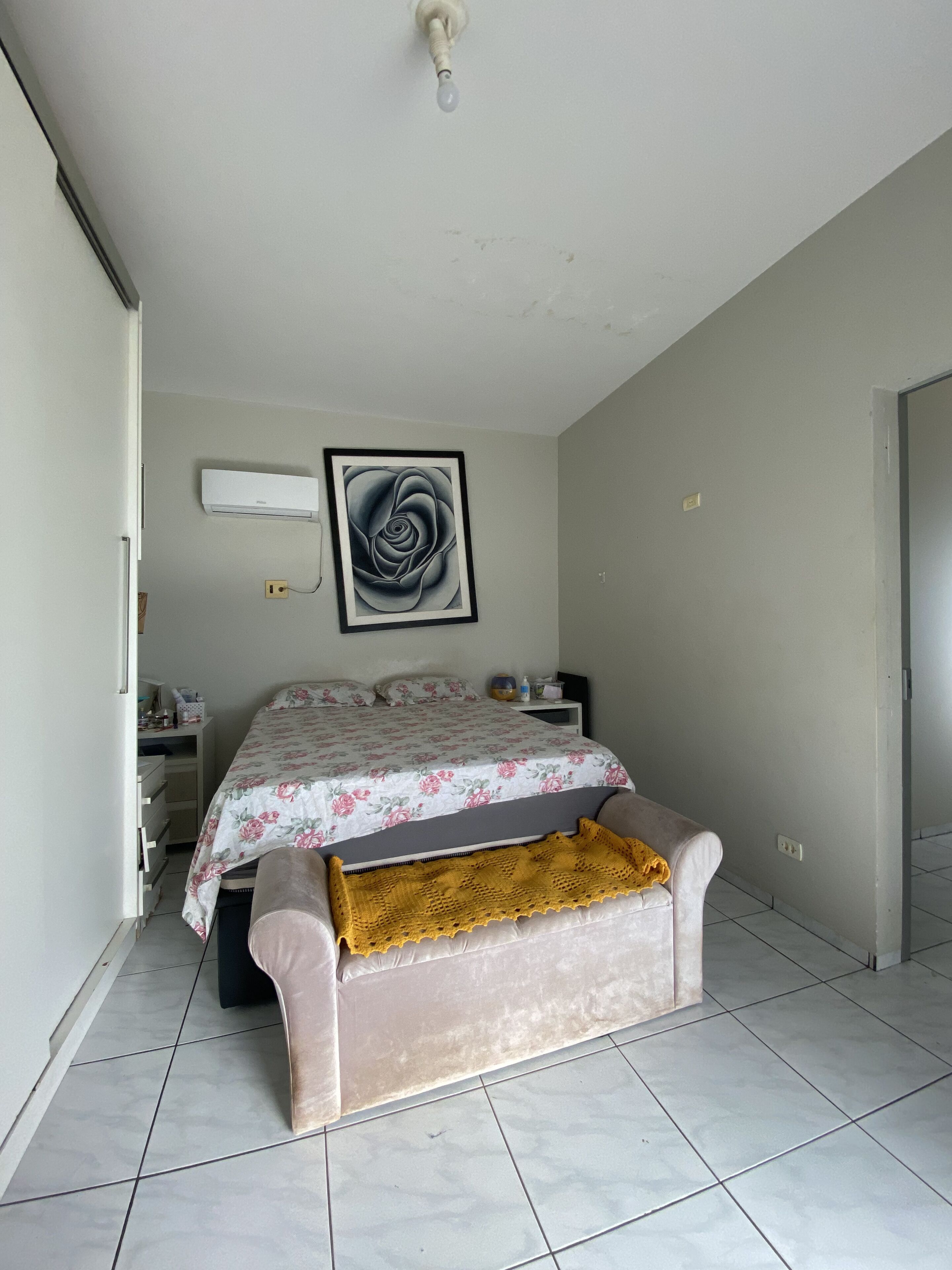 A bedroom with a bed, a bedside table, a nightstand, a wall-mounted air conditioner, and a framed artwork.