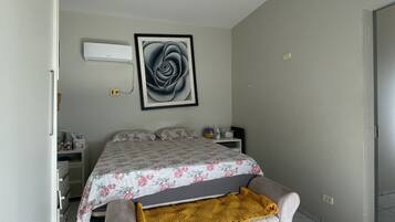A bedroom with a bed, a bedside table, a nightstand, a wall-mounted air conditioner, and a framed artwork.