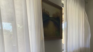Una habitación con un amplio ventanal cubierto por cortinas blancas translúcidas, un cuadro enmarcado en la pared, y dos jarrones negros con orquídeas blancas sobre una consola de madera oscura.