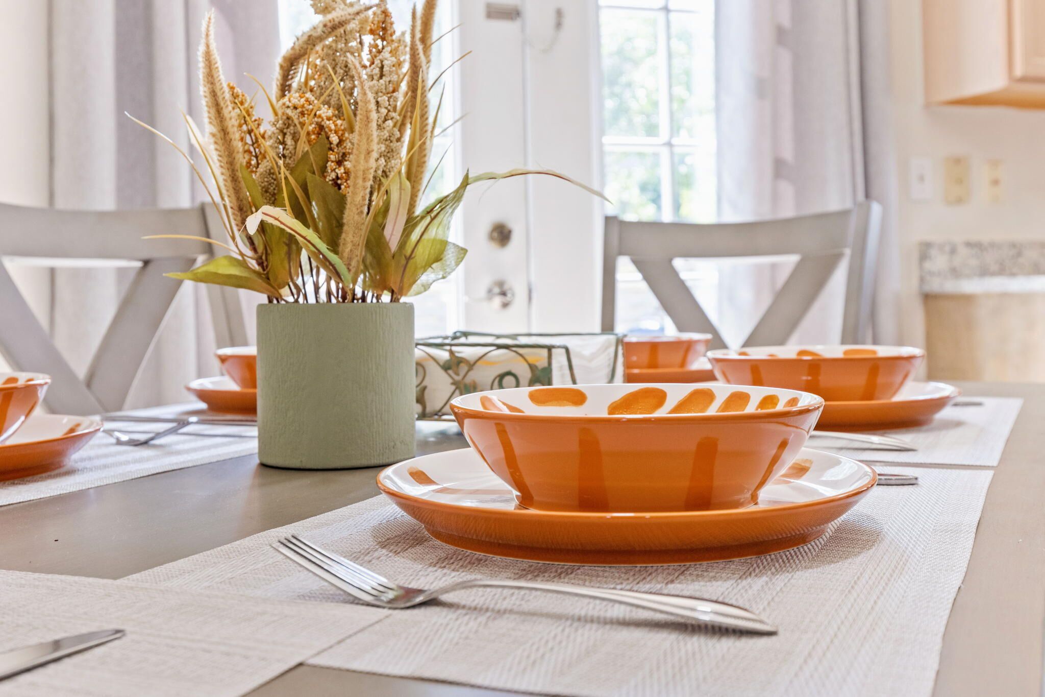 Une table à manger dressée avec des assiettes et des bols orange, un vase contenant des plantes séchées, et une porte blanche en arrière-plan.