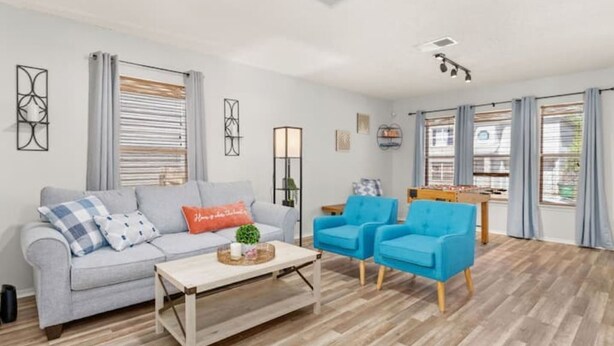 Een moderne woonkamer met een grijze bank, blauwe fauteuils, een houten salontafel en een tafelvoetbaltafel.